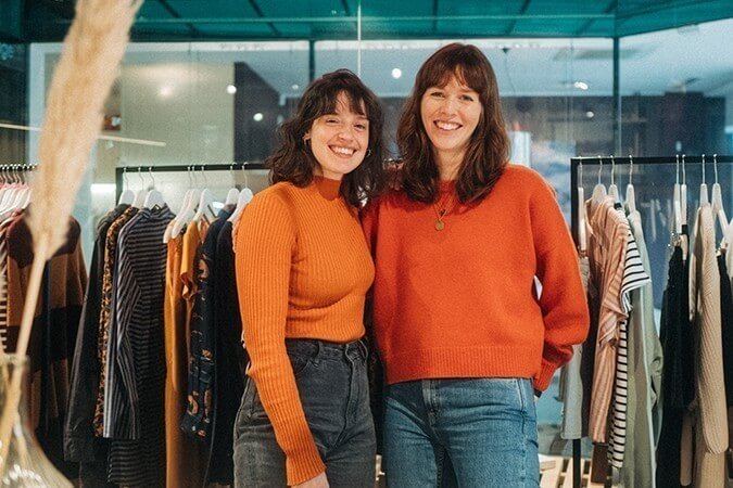Lächelnde Mitarbeiter des glore Ladens mit orangenen Oberteilen und Jeans stehen vor Kleiderstangen mit verschiedenen Kleidungstücken