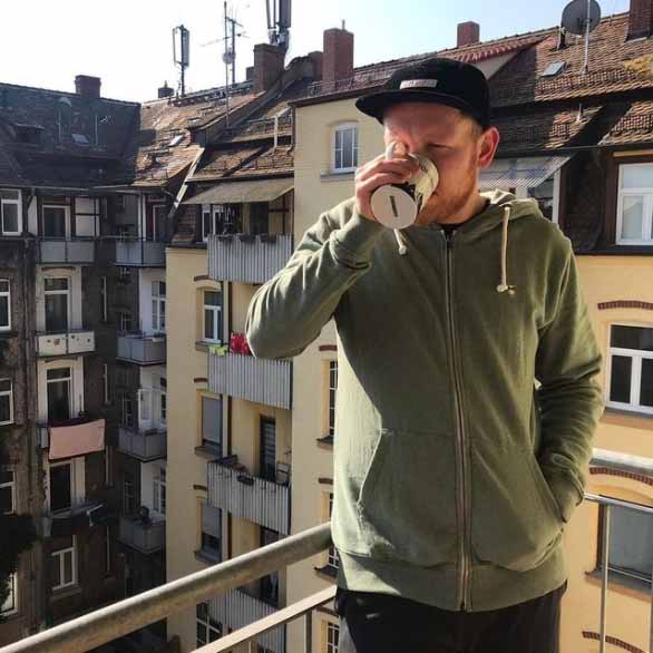 Man trinkt Kaffee auf Balkon und trägt eine schwarze Cap und grünen Sweatjacke