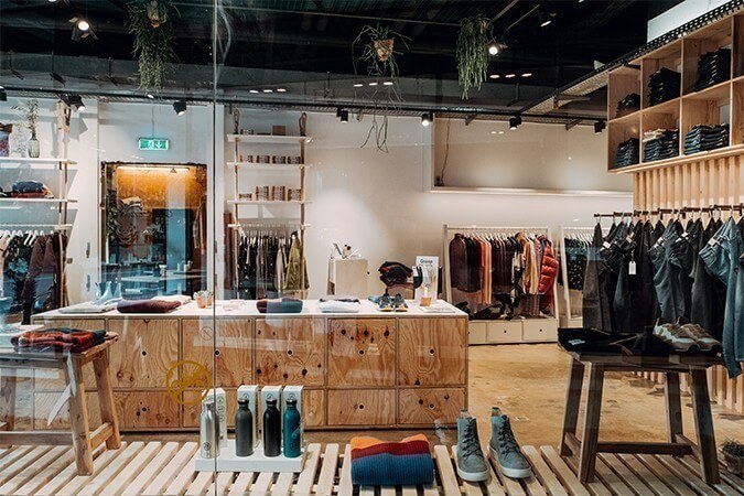 Einblick in den Laden durch Fensterscheibe mit Blick auf nachhaltiges Jeansregal, Holzblock mit Auslage und Kleiderstangen mit nachhaltigen Kleidungsstücken.