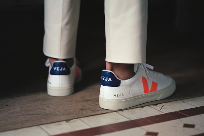 VEJA bei glore, Person mit heller Stoffhose trägt nachhaltige, weiße Sneaker mit farblich abgesetztem Logo in neon Orange und Fersenkappe in blau